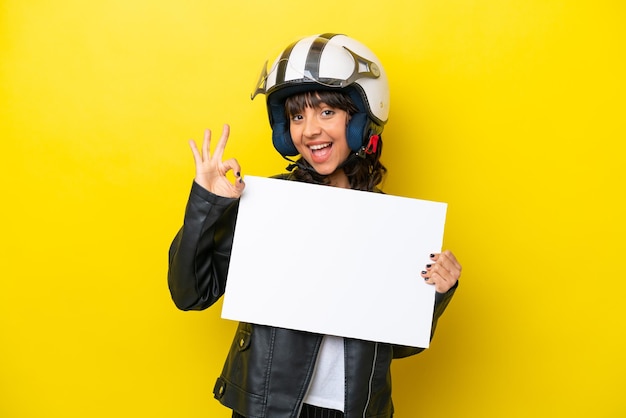 Joven latina con un casco de motocicleta aislado de fondo amarillo sosteniendo un cartel vacío y haciendo el signo de OK