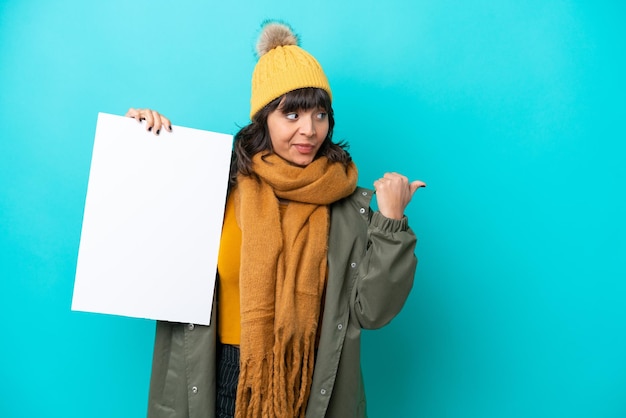 Joven latina con chaqueta de invierno aislada de fondo azul sosteniendo un cartel vacío y apuntando hacia el lado