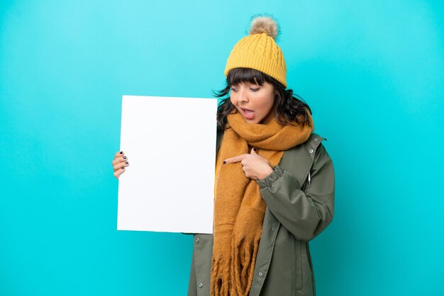 Joven latina con chaqueta de invierno aislada de fondo azul sosteniendo un cartel vacío con expresión feliz y apuntándolo