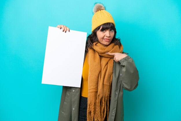 Joven latina con chaqueta de invierno aislada de fondo azul sosteniendo un cartel vacío con expresión feliz y apuntándolo