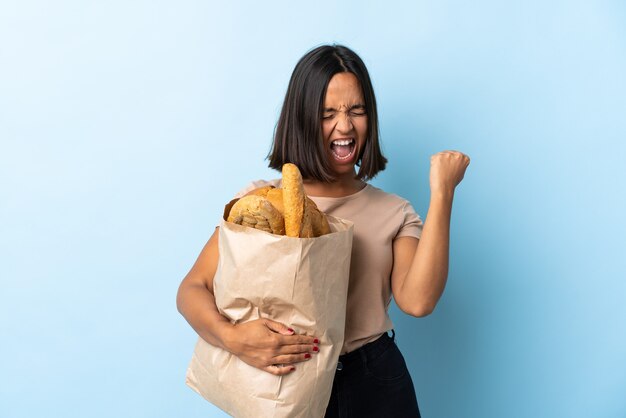 Joven latina comprando algunos panes aislados en azul celebrando una victoria