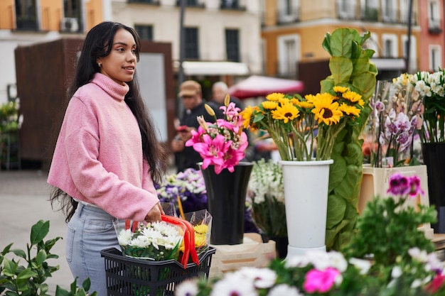 Una joven latina comprando plantas con una canasta llena de ellas en una floristería callejera