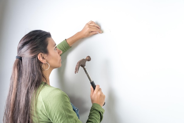 Joven latina confundida con clavo y martillo mirando una pared blanca nueva casa trabajo en casa mujer trabajadora