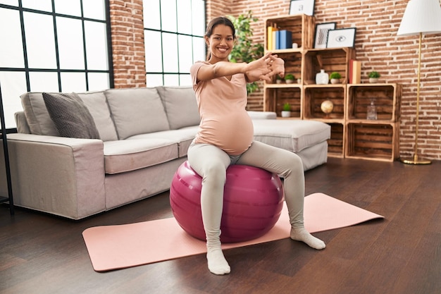 Joven latina embarazada estirando usando fit ball en casa