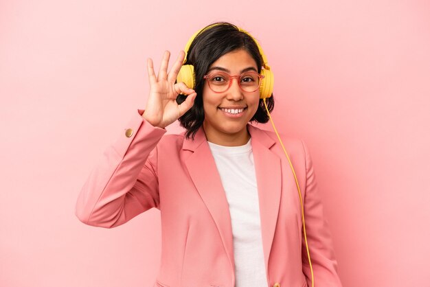 Joven latina escuchando música aislada sobre fondo rosa alegre y confiado mostrando gesto ok.