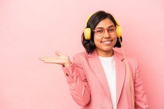 Joven latina escuchando música aislada sobre fondo rosa mostrando un espacio de copia en una palma y sosteniendo otra mano en la cintura.