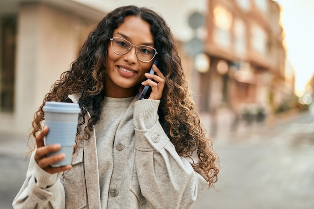 Joven latina hablando por teléfono inteligente y tomando café en la ciudad.