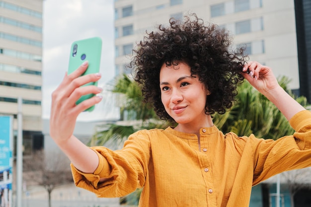 Foto joven latina haciendo un retrato de selfie con un teléfono celular tocando su cabello rizado feliz hismanic girl tomando una foto con un dispositivo de teléfono inteligente