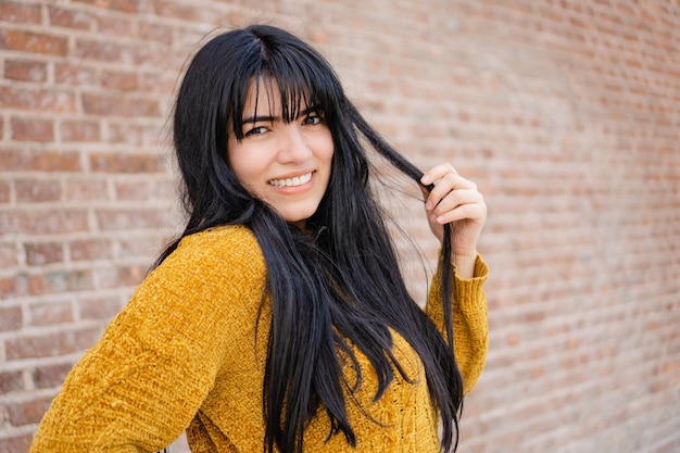 Joven latina hispana y caucásica con cabello lacio sonriendo mientras sostiene su cabello Fondo de ladrillo Copiar espacio