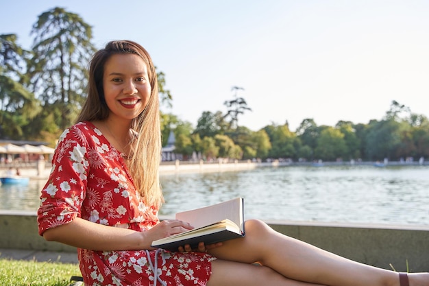 Joven latina leyendo un libro sentada en el césped a la orilla de un lago mirando a la cámara