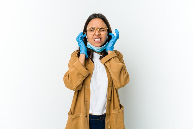 Joven latina con una máscara para protegerse del covid aislado en blanco coning orejas con las manos.