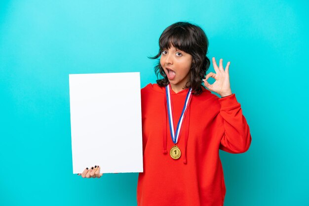 Joven latina con medallas aisladas de fondo azul sosteniendo un cartel vacío y haciendo el signo de OK