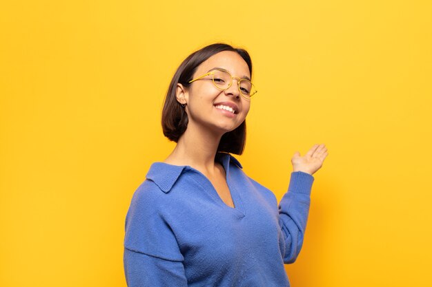 Joven latina que se siente feliz y alegre, sonriendo y dándote la bienvenida, invitándote con un gesto amistoso