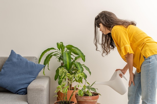 Joven latina regando las plantas con una regadera blanca en la sala de estar, vestida informalmente, con una blusa amarilla y un jean azul