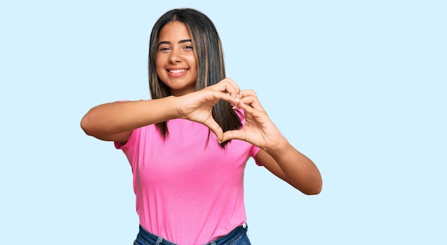 Joven latina con ropa informal sonriendo enamorada haciendo forma de símbolo de corazón con las manos. concepto romántico.