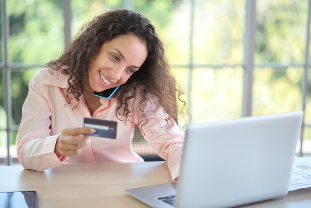 Una joven latina se sienta en su casa con una tarjeta de crédito para comprar en línea con una computadora.