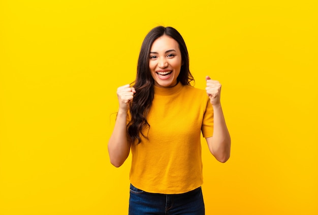 Joven latina se siente sorprendida, emocionada y feliz, riendo y celebrando el éxito, diciendo ¡guau! en pared plana