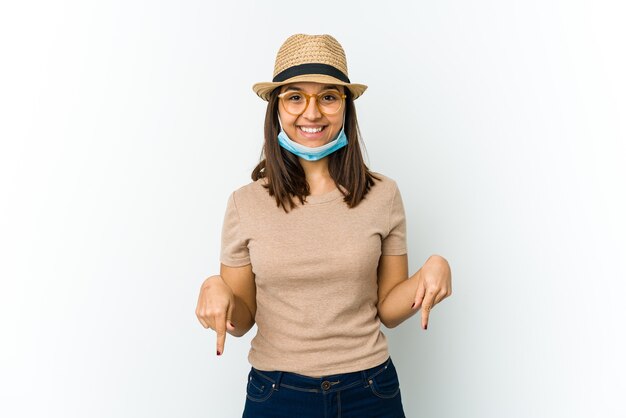 Joven latina con sombrero y máscara para protegerse del covid aislado en puntos blancos hacia abajo con los dedos, sentimiento positivo.