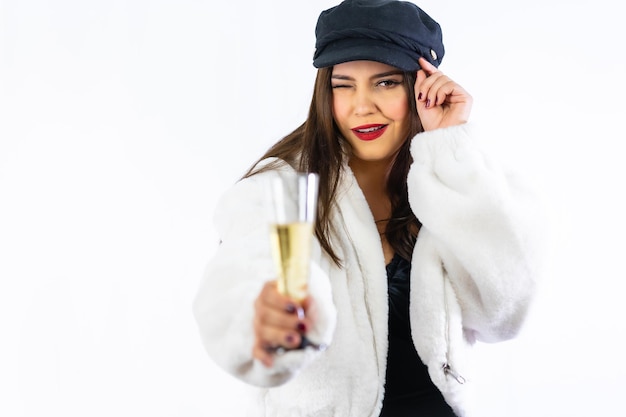 Joven latina con sombrero negro celebrando la víspera de año nuevo sobre un fondo blanco. Retrato guiñando un ojo con una copa de champán