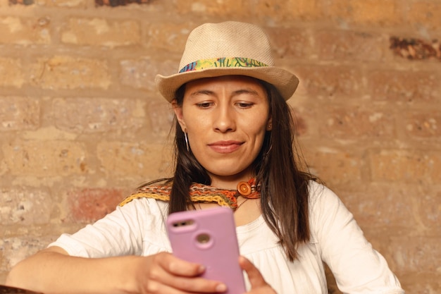 Joven latina con sombrero sentada en una silla de madera mirando su teléfono móvil contra una pared de barro