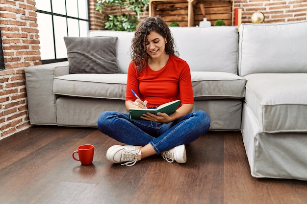 Joven latina sonriendo confiada escribiendo en un libro en casa