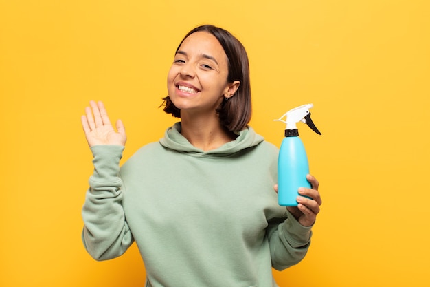 Joven latina sonriendo feliz y alegremente, saludando con la mano, dándote la bienvenida y saludándote, o despidiéndote