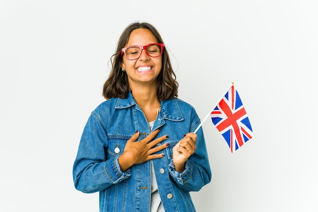 Joven latina sosteniendo una bandera inglesa se ríe alegremente y se divierte manteniendo las manos sobre el estómago.