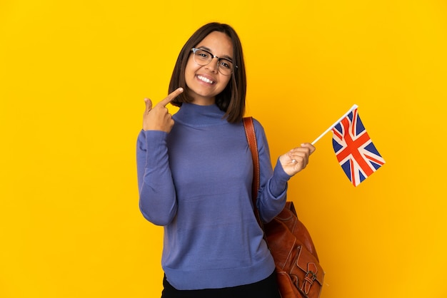 Joven latina sosteniendo una bandera de Reino Unido aislada en la pared amarilla dando un pulgar hacia arriba gesto