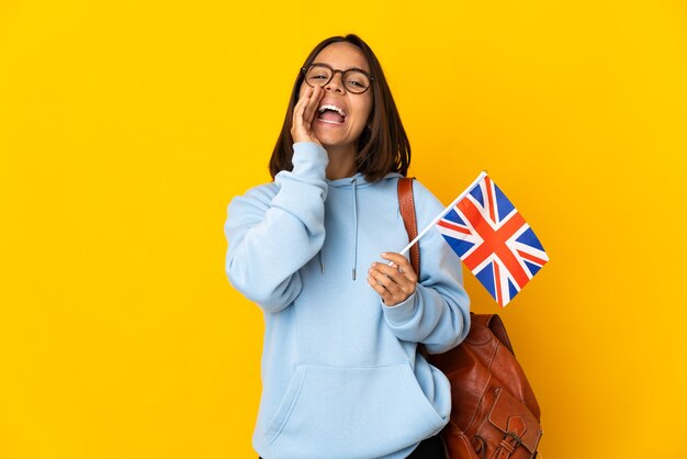 Joven latina sosteniendo una bandera del Reino Unido aislada en la pared amarilla gritando y anunciando algo