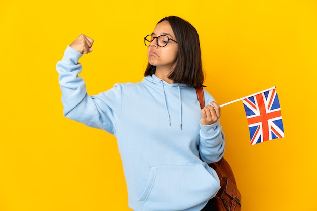 Joven latina sosteniendo una bandera de Reino Unido aislada en la pared amarilla haciendo un gesto fuerte