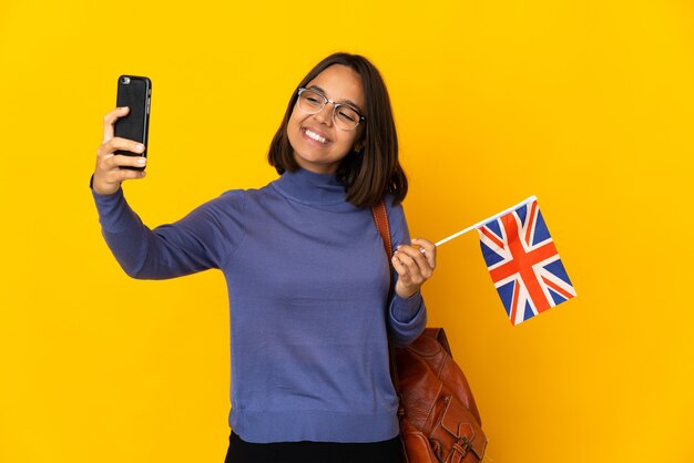 Joven latina sosteniendo una bandera del Reino Unido aislada en la pared amarilla haciendo un selfie