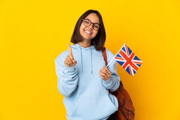 Joven latina sosteniendo una bandera del Reino Unido aislada en la pared amarilla mostrando y levantando un dedo