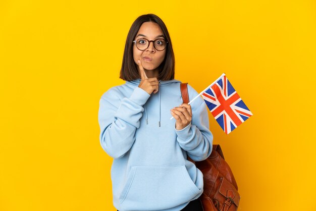 Joven latina sosteniendo una bandera del Reino Unido aislada en la pared amarilla y pensando