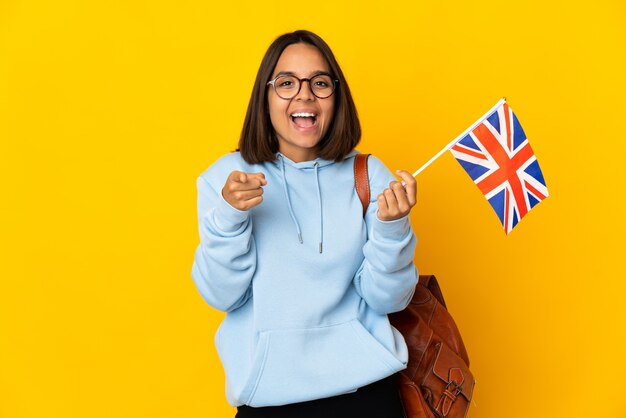 Joven latina sosteniendo una bandera del Reino Unido aislada en la pared amarilla sorprendida y apuntando hacia el frente