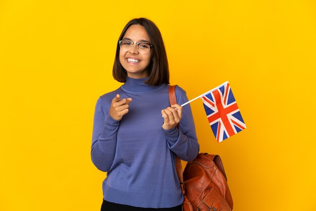 Joven latina sosteniendo una bandera del Reino Unido aislada en la pared amarilla sorprendida y apuntando hacia el frente