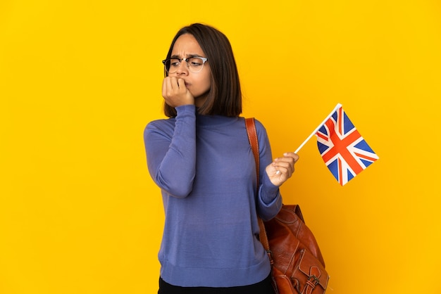 Joven latina sosteniendo una bandera del Reino Unido aislada en la pared amarilla teniendo dudas