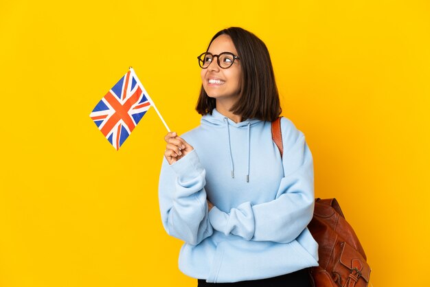 Joven latina sosteniendo una bandera del Reino Unido aislada sobre fondo amarillo feliz y sonriente