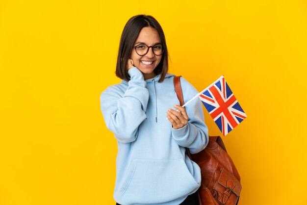 Joven latina sosteniendo una bandera del Reino Unido aislado en la pared amarilla riendo