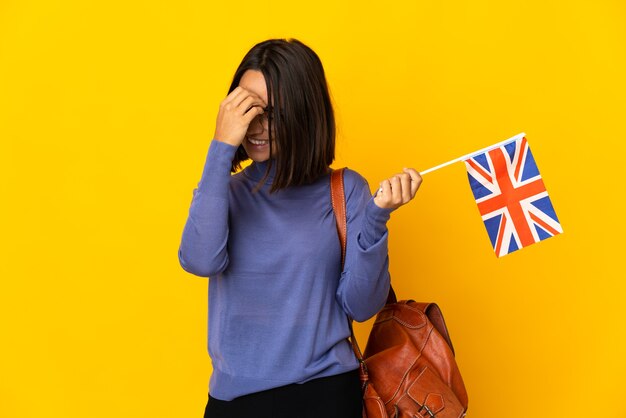 Joven latina sosteniendo una bandera del Reino Unido aislado en la pared amarilla riendo