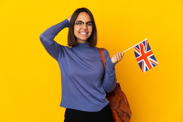 Joven latina sosteniendo una bandera del Reino Unido aislado sobre fondo amarillo riendo