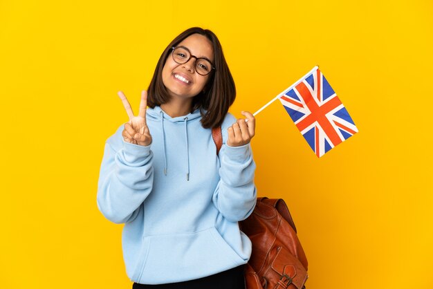 Joven latina sosteniendo una bandera del Reino Unido aislado sobre fondo amarillo sonriendo y mostrando el signo de la victoria