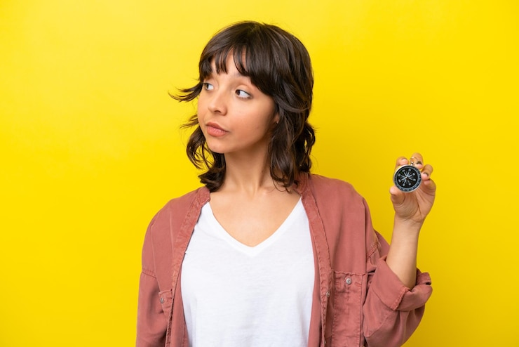 Foto joven latina sosteniendo una brújula aislada en un fondo amarillo mirando hacia un lado
