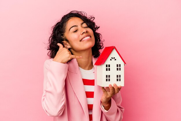 Foto joven latina sosteniendo una casa de juguete aislada sobre fondo rosa mostrando un gesto de llamada de teléfono móvil con los dedos.