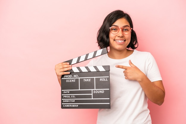 Joven latina sosteniendo claqueta aislada sobre fondo rosa sonriendo y apuntando a un lado, mostrando algo en el espacio en blanco.