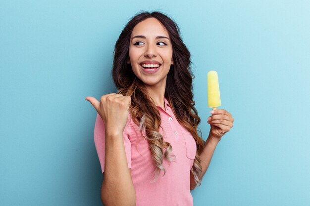Joven latina sosteniendo helado aislado sobre fondo azul apunta con el dedo pulgar lejos, riendo y despreocupado.
