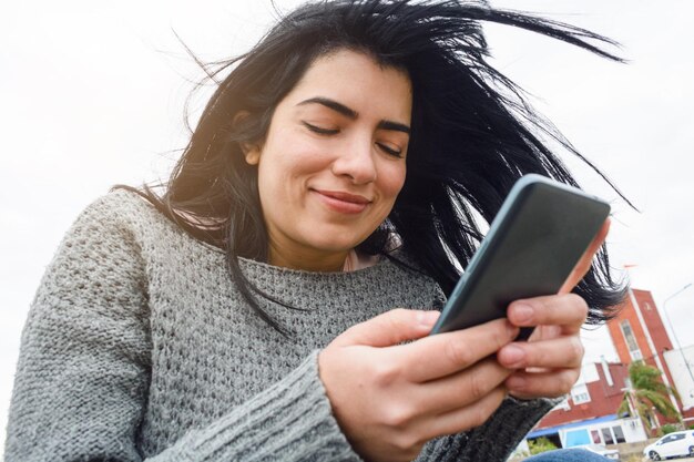 joven latina usando el teléfono tranquila sentada al aire libre en un día nublado y frío