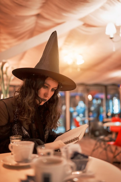 Joven latina vestida de bruja sentada y leyendo un cuaderno en un cafex9