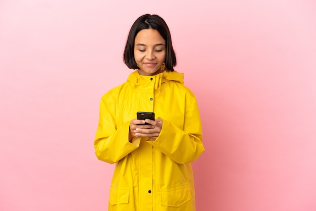 Joven latina vistiendo un abrigo impermeable sobre antecedentes aislados enviando un mensaje con el móvil