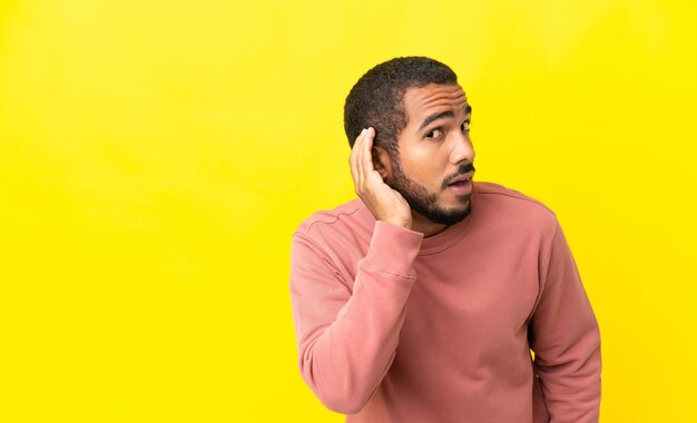 Joven latino aislado de fondo amarillo escuchando algo poniendo la mano en la oreja