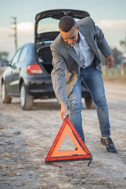 Joven latino colocando un triángulo de advertencia detrás de un auto averiado